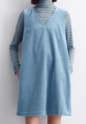 Vrouw draagt een lichtblauwe mouwloze corduroy jurk over een blauw-beige gestreepte coltrui met handen in de zakken.