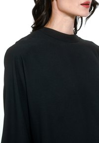 Črna bluzica iz gladkega materiala, z visokim ovratnikom in ohlapnimi rokavi, z mehko teksturo in minimalnimi oblikovalskimi podrobnostmi.