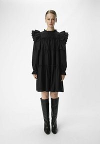 Schwarzes Kleid mit puffärmeligen Ärmeln, gerafften Stufen, rüschigem Ausschnitt und langen Ärmeln, kombiniert mit knielangen schwarzen Lederstiefeln.