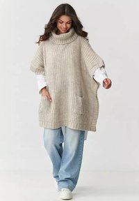 Kobieta ubrana w oversize'owy beżowy sweter z golfem i kieszeniami z przodu, jasnoniebieskie dżinsy i białe trampki, spacerująca z uśmiechem.