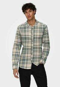 Camisa de cuadros en colores verde y crema, hecha de tela suave. Presenta un cierre frontal con botones, mangas largas y botones negros.