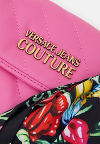 Rosa läderclutch med kviltad textur och guld "VERSACE JEANS COUTURE"-logotyp. Blommönstrat tyg syns från sidan.