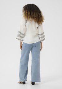 Blusa bianca ricamata con maniche ampie e dettagli neri, abbinata a jeans a gamba larga di colore blu chiaro e sandali con tacco medio neri.