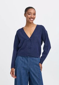 Cardigan a maglia blu navy con scollatura a V profonda, chiusura frontale con tre bottoni e maniche lunghe, abbinato a pantaloni in denim.