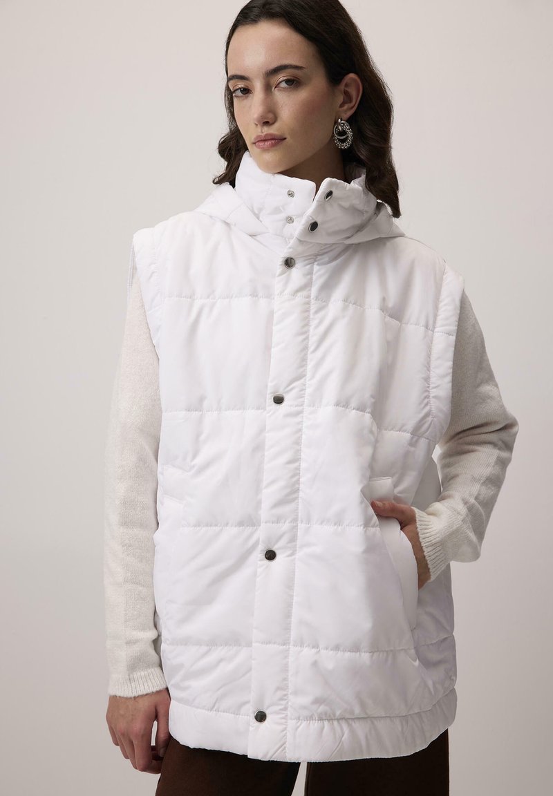 Bijela bezrukavna puffer prsluk s visokim ovratnikom, dugmadi na pritiskanje i bočnim džepovima. Tekstuirana tkanina s quilted uzorkom, složena preko kremaste bluze.