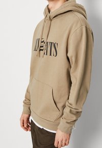 Beżowa bluza z kapturem z przednią kieszenią kangurka, ściągaczami na mankietach i oversize'owym krojem. Na piersi znajduje się wyrazisty czarny napis.