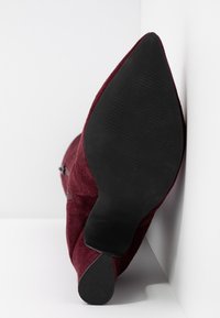 Bota de tacón alto de ante burdeos con punta afilada y suela de goma negra. El tacón es delgado y la puntera está elegantemente estrechada.