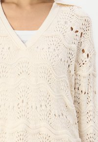 Cremig gestrickter Pullover mit V-Ausschnitt, der ein Wellenmuster und eine offen gearbeitete Gestaltung aufweist. Texturiertes Material und lockere Passform, ideal zum Schichten.