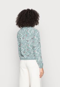 Vrouw met krullend haar, gekleed in een lichtblauwe lange mouwtop met een patroon van vogels en bladeren, en een witte broek, met haar rug naar de camera gekeerd.