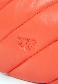 Koralo odinė rankinė su siuvinėta dizainu, turinti įstrižai siūtas detales ir subtilų logotipą iškilusį ant paviršiaus. Matoma lygi tekstūra.