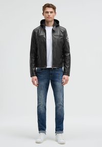 Schwarze Lederjacke mit Kapuze, Frontreißverschluss, zwei Brusttaschen, über einem weißen T-Shirt getragen, kombiniert mit blauen Jeans und weißen Sneakern.