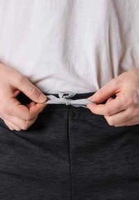 Grå midjeband på mörka träningsshorts, med en dragsko som knyts, händer placerade vid midjan och en helt vit topp synlig ovanför.