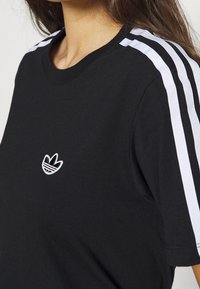 Černé bavlněné tričko s bílým designem tří pruhů na rameni a vyšitým logem na hrudi. Kulatý výstřih a krátké rukávy.