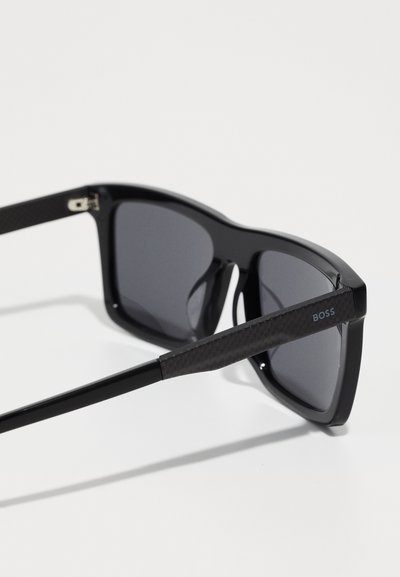 Lunettes de soleil noires avec des montures carrées, des verres foncés et des branches noires texturées arborant un petit logo bleu "BOSS".