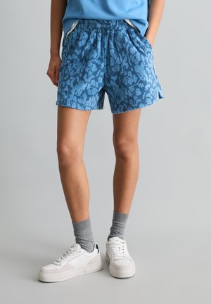 Osoba nosząca niebieskie spodenki z froty w wzory, szare skarpetki, białe trampki oraz niebieską koszulę wpuszczoną do spodni, stojąca na szarej podłodze.