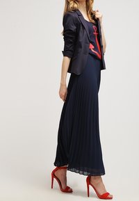 Mörkblå plisserad maxikjol tillsammans med en figurnära mörkblå topp med en röd ankardesign och en strukturerad svart blazer. Bärs med röda högklackade skor.