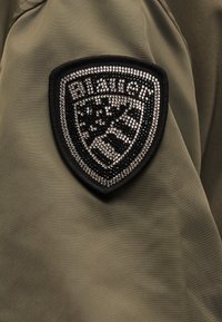 Μαύρο και ασημί έμβλημα ασπίδας με στρας σε ελιά πράσινο ύφασμα, που περιλαμβάνει τη λέξη "Blauer" και αφηρημένο ριγωτό σχέδιο.