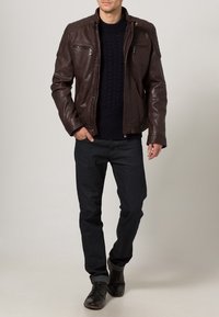 Tmavě hnědá kožená bunda se zipy, námořnicky modrý svetr s copánkovým vzorem, tmavé džíny a černé šněrovací boty. Model stojící před neutrálním pozadím.