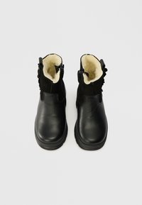 Czarne skórzane botki za kostkę z zamszowym wykończeniem, z kremowym futerkiem i zamkami po bokach, pokazane z góry na białym tle.