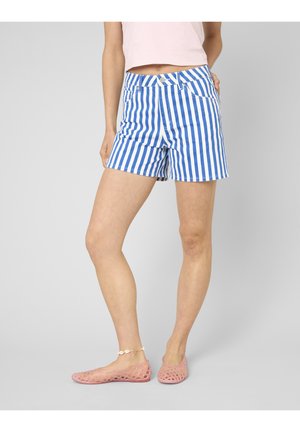 Femme portant un short rayé vertical bleu et blanc, un crop top rose clair, des ballerines en mesh roses et un bracelet de cheville en coquillages, debout avec une main dans la poche.