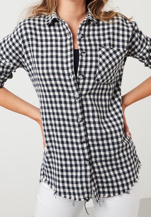 Kobieta w czarno-białej koszuli w kratę zapinanej na guziki, z podwiniętymi rękawami i postrzępionym dołem, w połączeniu z białymi spodniami.