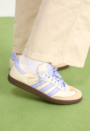 Baskets Adidas Samba beige et lavande portées avec des chaussettes blanches et un pantalon beige sur un sol recouvert de moquette verte.