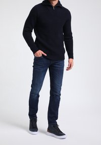 Pull en maille côtelée bleu marine avec col à demi-fermeture éclair, associé à un jean bleu foncé et des baskets montantes noires avec semelles blanches.