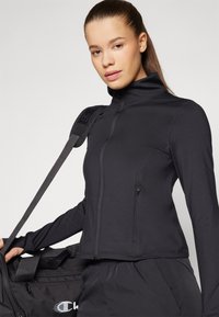 Musta urheiluuntuvä jacket, jossa on korkea kaulus, vetoketjusulku ja muotoiltu malli. Päällä on vastaavat alaosat, joissa on logo takana.