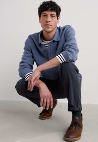 Sininen pitkähihaisen nappipaitapusero, jossa on raidalliset hihansuut, puettuna raidallisen paidan päälle. Tummat housut ja ruskeat nahkakengät täydentävät asun.