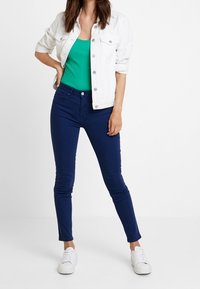 Chaqueta de mezclilla blanca, camiseta sin mangas de canalé verde y jeans ajustados azul marino. Completado con zapatillas blancas, creando un atuendo casual.