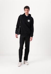 Hanorac negru cu buzunar frontal, logo brodat, asociat cu pantaloni negri și adidași albi. Material neted, croială lejeră, elemente de design minime.