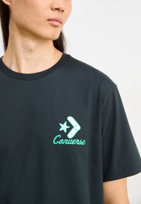 Camiseta de algodón negra con un logo de Converse en turquesa, que incluye una estrella y un diseño de chevron. Cuello redondo estándar y mangas cortas.