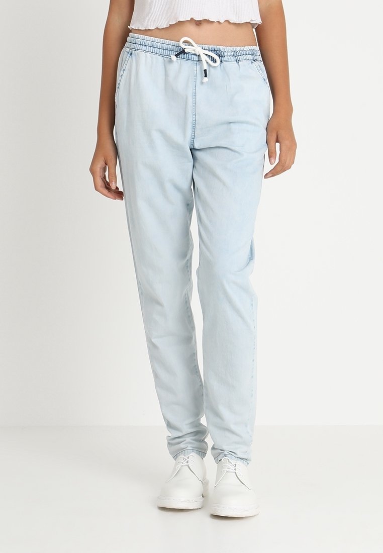 Personne portant un pantalon bleu clair à cordon avec poches latérales et des chaussures blanches à lacets, debout devant un fond uni.
