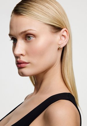 Złota kolczyk w kształcie obręczy na modelce z prostymi blond włosami, ubranej w czarny top. Kolczyk ma gładki, zaokrąglony kształt.