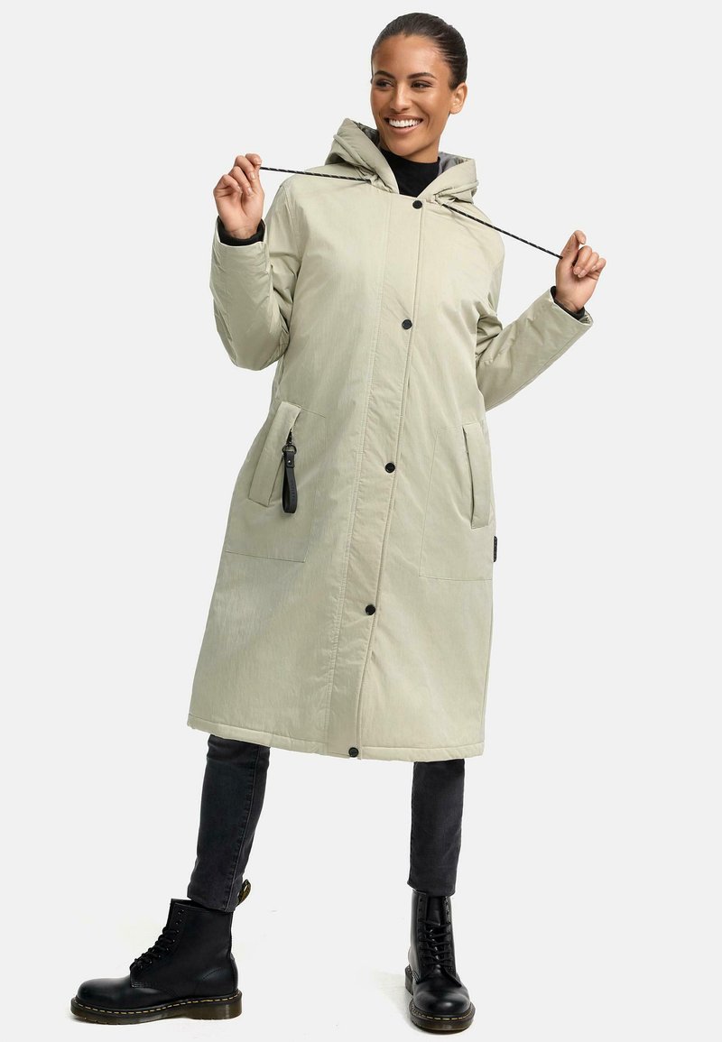 Lang, lichtbeige gewatteerde jas met een capuchon, voorzien van zwarte knopen en twee voorzakken. Draag het over een zwarte top en gecombineerd met zwarte veterschoenen.