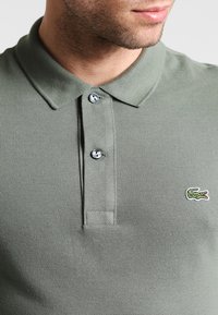 Polo vert olive, en tissu texturé. Il comporte un col, une patte de boutonnage à deux boutons et un petit logo brodé sur la poitrine.