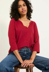 Bordeauxrotes langärmeliges Oberteil mit V-Ausschnitt und drei Knöpfen, kombiniert mit hochgeschnittenen blauen Jeans. Der Stoff sieht weich und mit einer glatten Textur aus.