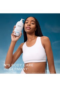 White bottle with pump dispenser, contains body lotion, minimalistic design. Model with long braided hair, wearing a white crop top, blue background.