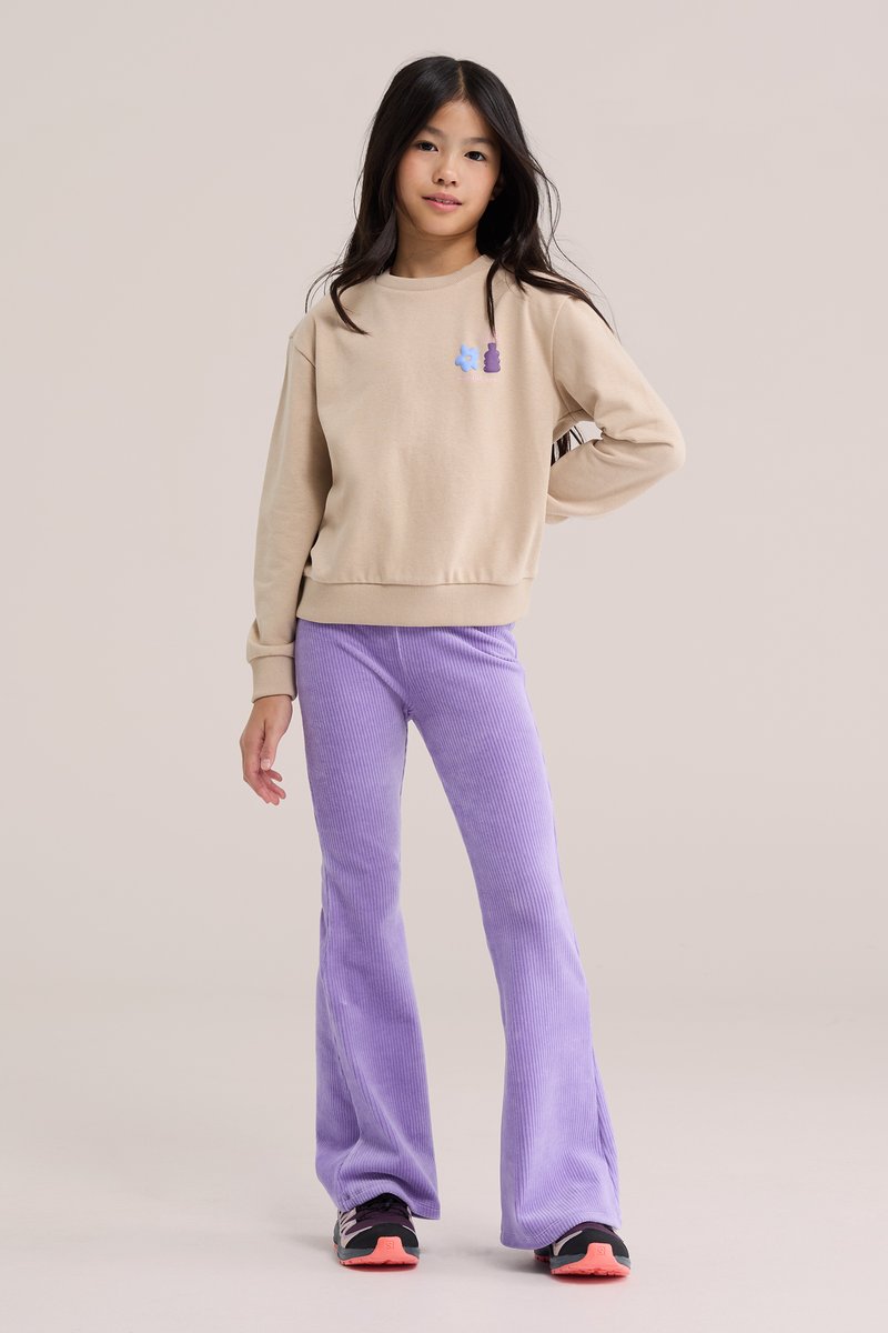Fille aux longs cheveux foncés, debout, main sur la hanche, portant un sweat-shirt beige et un pantalon évasé en velours côtelé violet avec des chaussures noires et roses.