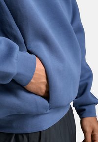 Eine Nahaufnahme einer Person in einem blauen Sweatshirt, mit einer Hand, die in die Fronttasche gesteckt ist, wobei die Stofftextur und der Schnitt hervorgehoben werden.