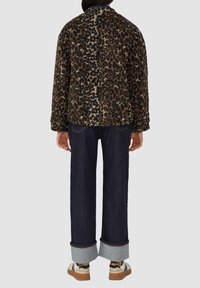 Persoon in een bruine en zwarte luipaardprintjacket, donkere opgerolde jeans en witte sneakers met rubberen zolen, met de rug naar de camera op een grijze achtergrond.