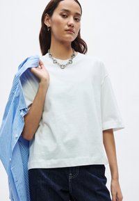 Balta oversize kokvilnas T-krekls ar īsām piedurknēm, kombinēts ar gaiši zilu svītrainu kreklu, sudraba ķēdes kaklarotu un tumšām džinsa biksēm.