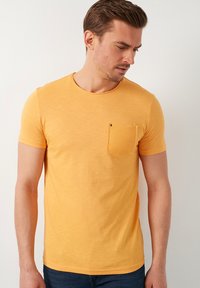 Žluté bavlněné tričko s kulatým výstřihem a krátkými rukávy. Obsahuje texturovanou kapsu na levém hrudníku s viditelným stehem.