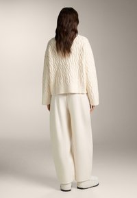 Prilagojen slonokoščeni pleteni pulover z vzorci tekstur, kombiniran z ohlapnimi slonokoščeno barvnimi hlačami. Model nosi bele čevlje, pogled od zadaj.
