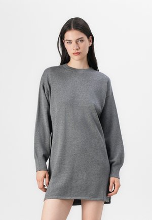 Jonge vrouw die een grijze, lange mouw sweaterdress modelleert tegen een eenvoudige witte achtergrond, staande met een neutrale uitdrukking.