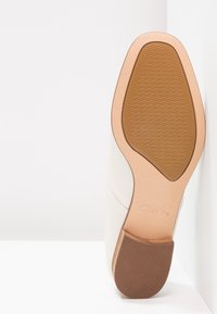 Talons blancs avec une semelle en caoutchouc beige, présentant un dessus lisse et une semelle en caoutchouc texturé en dessous. Design minimaliste et lignes épurées.