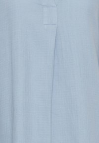 Světle modrý textil s jemnou mřížkovou strukturou, se v-neck výstřihem a krátkým detailním zapínáním. Měkká, tkaná textura.