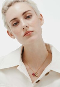 Collar de cadena de oro con un colgante en forma de luna creciente y una perla rosa. El modelo lleva una camisa blanca con cuello, lo que resalta la joyería.