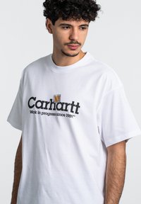Camiseta de algodón blanca con un estampado gráfico negro que presenta el texto "Carhartt WIP Work in Progress desde 1989" en la parte frontal. Mangas cortas.