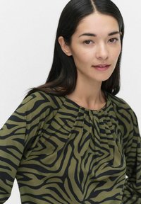 Armégrön blus med svart zebramönster, långa ärmar och rynkade veck vid halsen för textur och design.